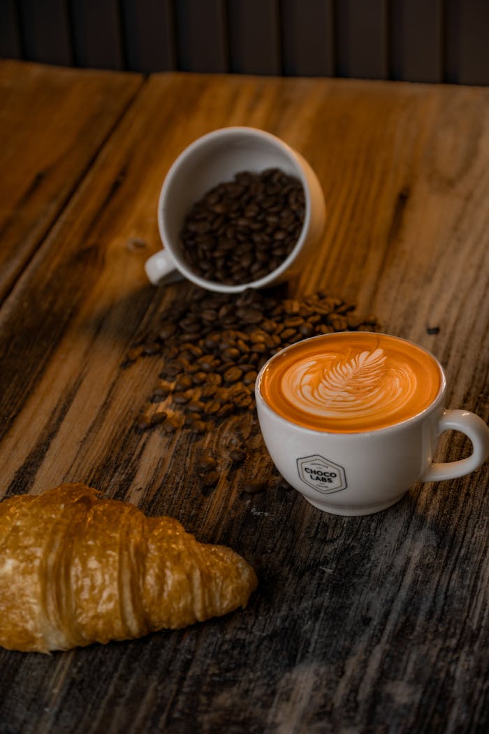 A cup of coffee with latte art, spilled coffee beans, and a croissant on a rustic wooden table.